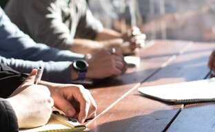 Personen sitzen um einen Tisch am Fenster und sprechen miteinander. Manche Personen haben einen Notizblock vor sich und schreiben etwas. Der Fokus liegt auf den Händen der Personen.
