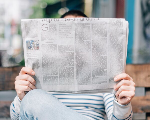 Eine Person sitzt auf einer Holzbank und wird dabei von ihrer Zeitung verdeckt