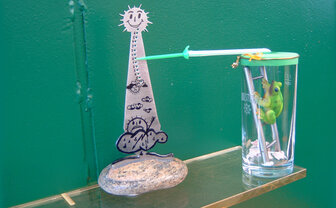 Eine gebastelte Wetterstation mit einem Plastikfrosch vor einem grünen Hintergrund.