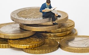 Eine kleine Miniatur-Figur sitzt mit verschränkten Beinen auf einem Stapel Euromünzen und liest eine Zeitung