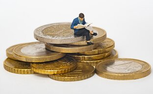 Eine kleine Miniatur-Figur sitzt mit verschränkten Beinen auf einem Stapel Euromünzen und liest eine Zeitung