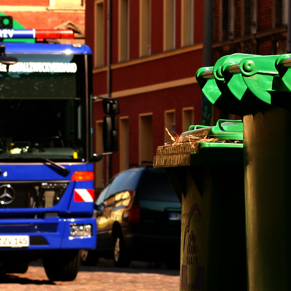 Mülltonnen stehen am vorderen linken Rand, während ein ein Müllauto im Hintergrund die andere Mülltonnen abholt