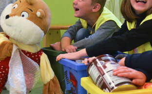 Drei Kinder sitzen hinter 3 kleinen Plastik-Abfalltonnen in grün, blau und gelb. Sie tragen gelbe Warnwesten. Vor ihnen sitzt eine Erzieherin mit Freddi dem Hamster im Arm.