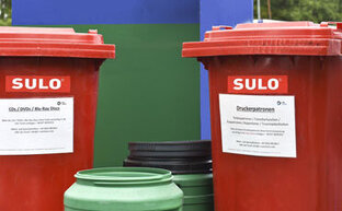 Zwei rote Mülltonnen für CDs und Toner stehen bei anderen Mülltonnen