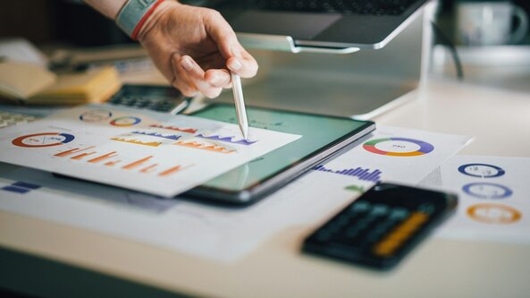 Eine Person tippt mit einem Stift auf einem Blatt Papier mit Statistiken. Das Blatt liegt auf einem Tablet mit weiteren Statistiken