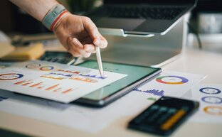 Eine Person tippt mit einem Stift auf einem Blatt Papier mit Statistiken. Das Blatt liegt auf einem Tablet mit weiteren Statistiken